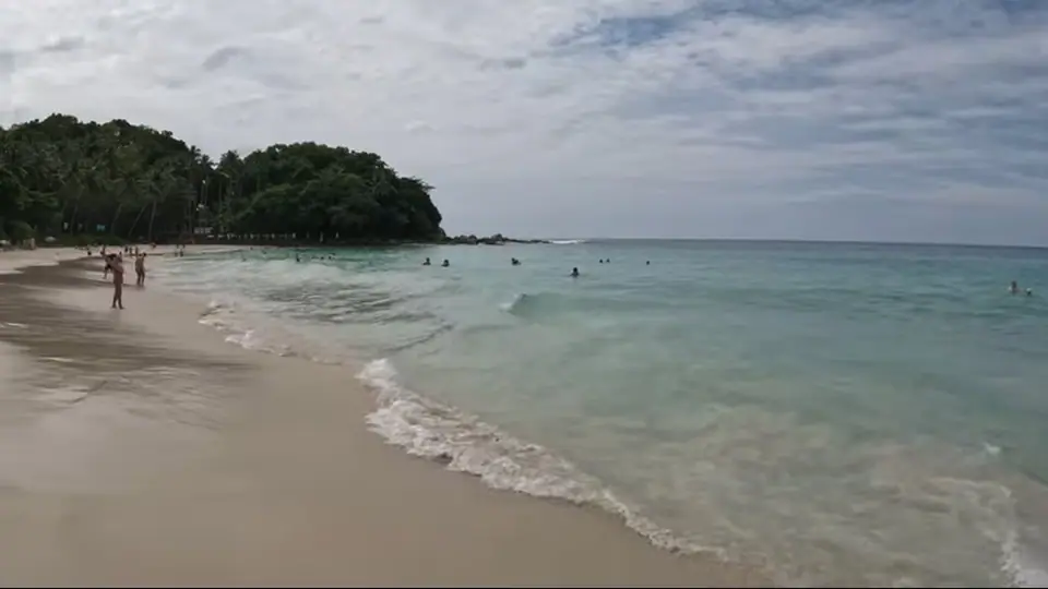 Calm translucent water meets pale sand with distant swimmers.