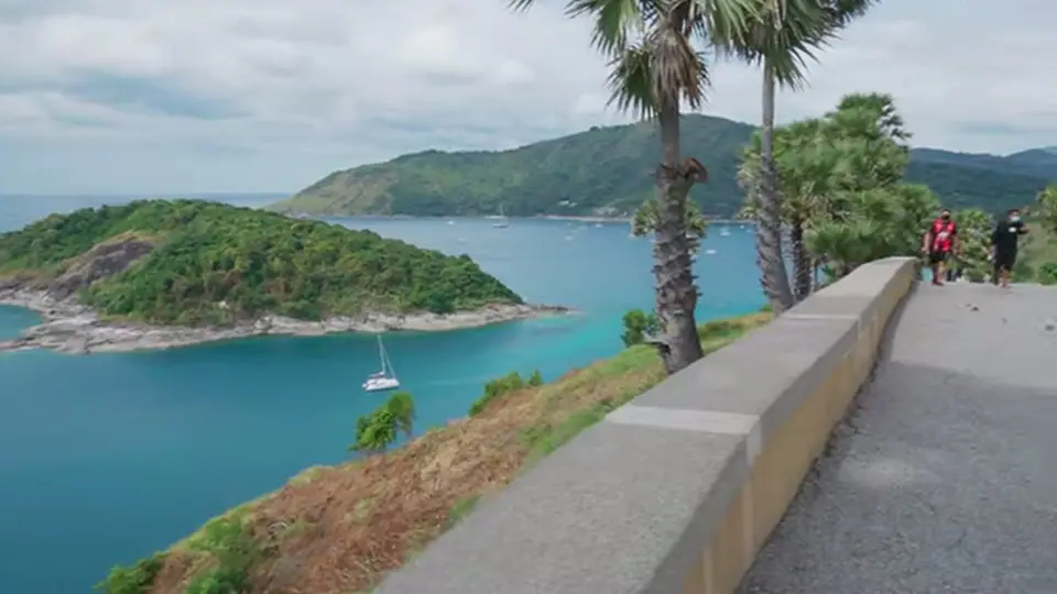 Coastal road above turquoise bay, palms, and a small sailboat.